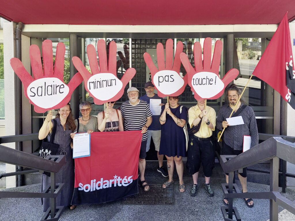 8 militant.e.s de solidaritéS avec deux drapeaux du parti ainsi que 4 pancartes en forme de mains rouges avec le slogan "salaire minimum pas touche !" (un mot par pancarte) devant les locaux de la CNCI